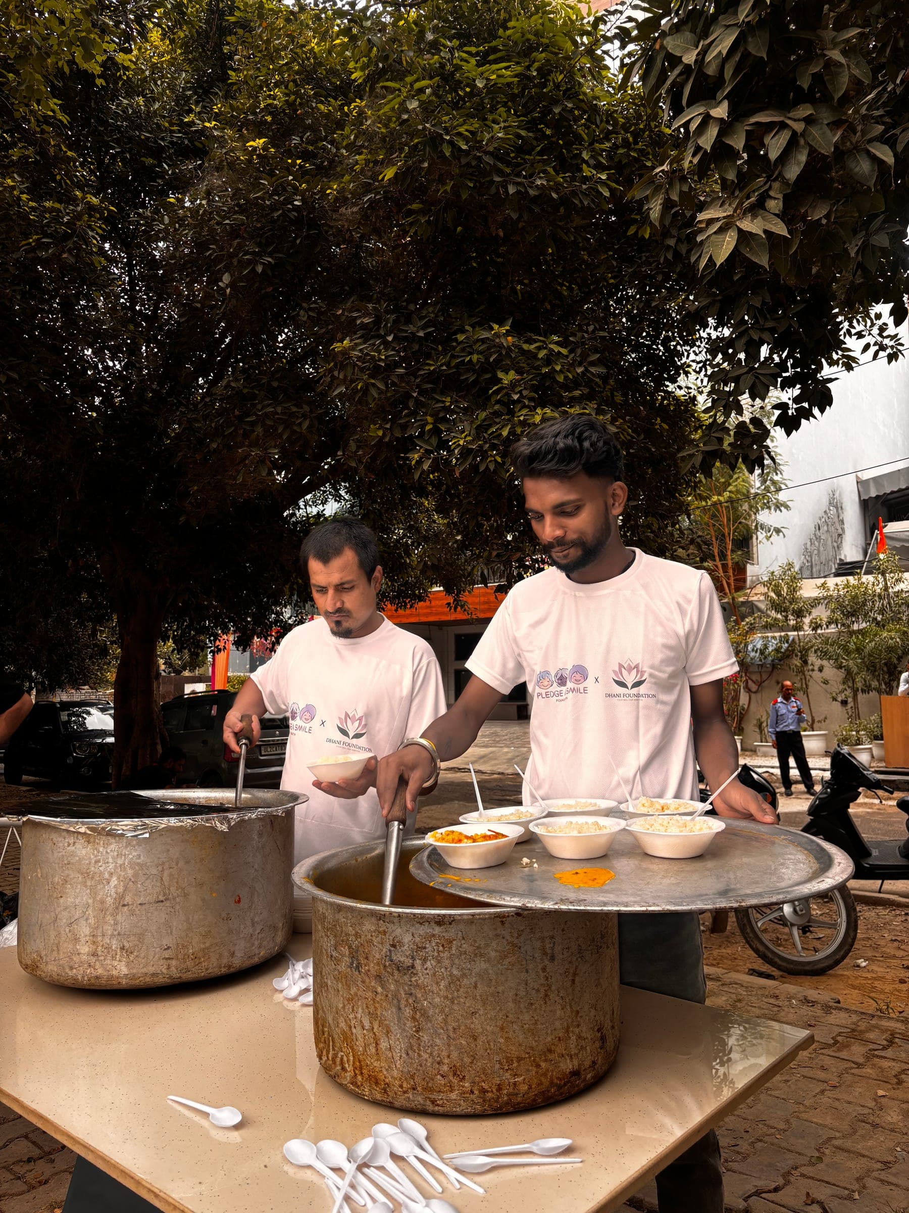 Two Pledge A Smile x Dhani Foundation volunteers plate rice and curry from large pots under a tree — the weekly Monday food drive.