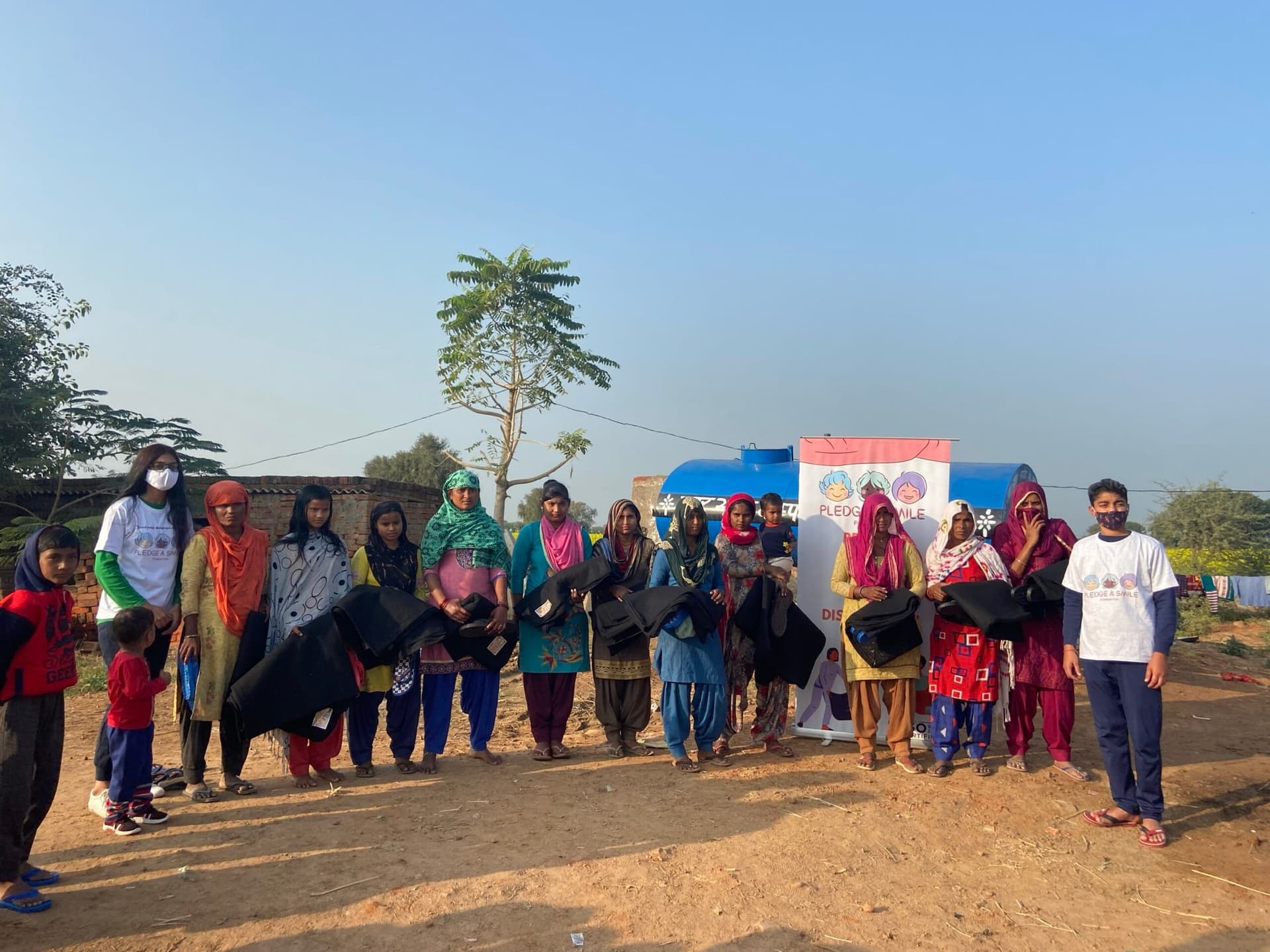 Winter-relief distribution at a village brick-kiln settlement in golden morning light; wide composition with a huge clean sky for headline text, real recipients holding blankets, PAS banner subtly in frame. Sharp 2560px file that survives a full-bleed crop.