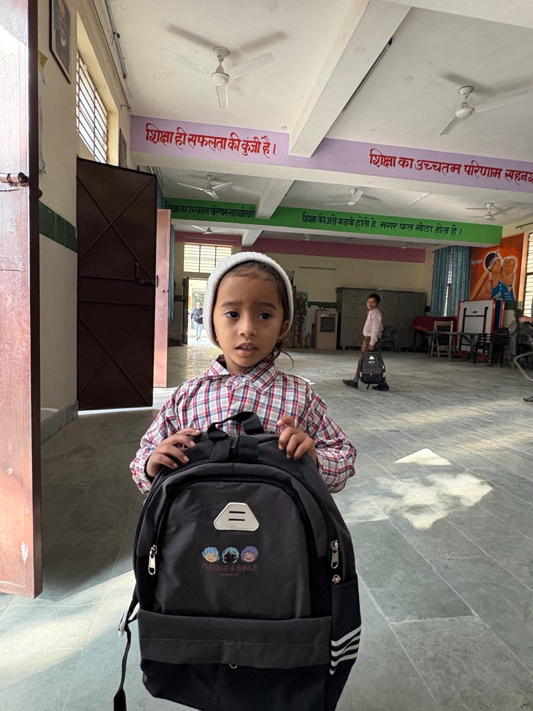 A small girl in a white knit cap presents her new backpack in a school corridor