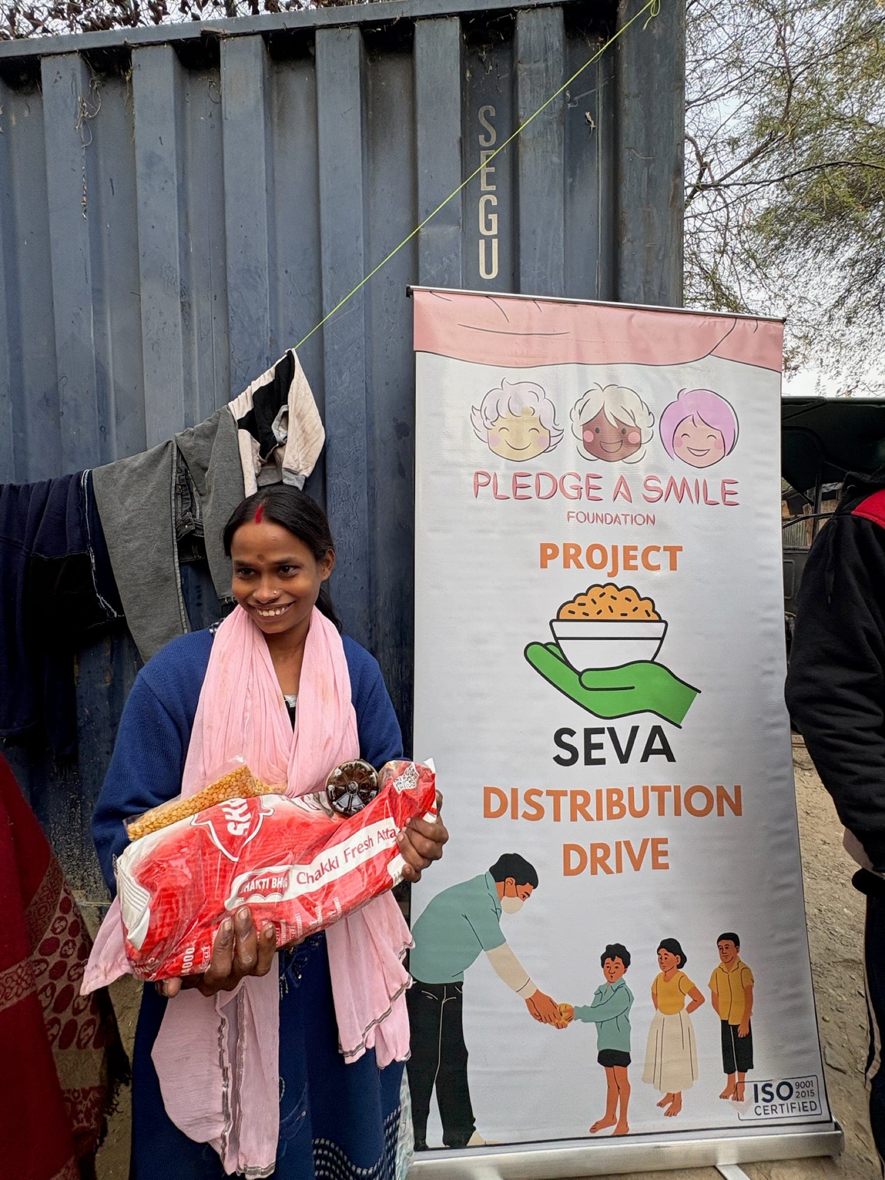 A woman in a pink shawl smiles beside the Seva banner, ration kit in hand