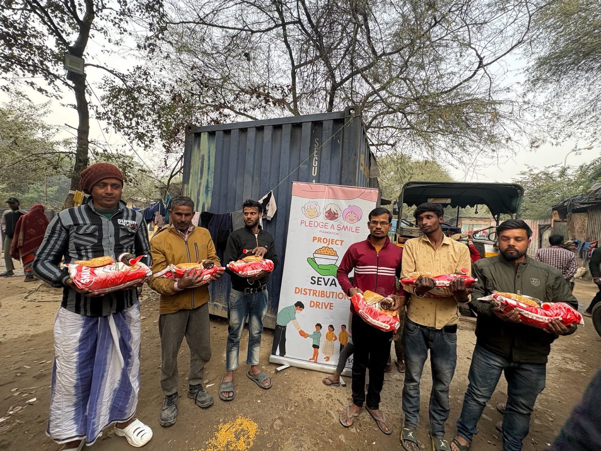 Wide scene: six men holding ration kits with SEVA banner at a labor-camp settlement, winter light