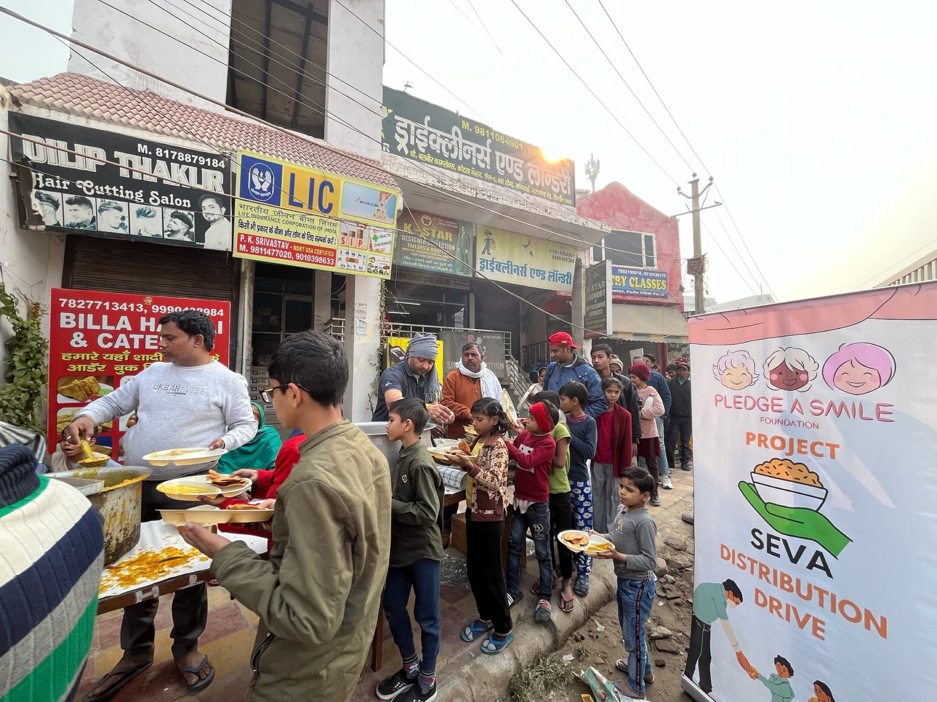 Wide winter street scene of the hot-food distribution queue on a Delhi-NCR street, the Project Seva banner anchoring the right edge.