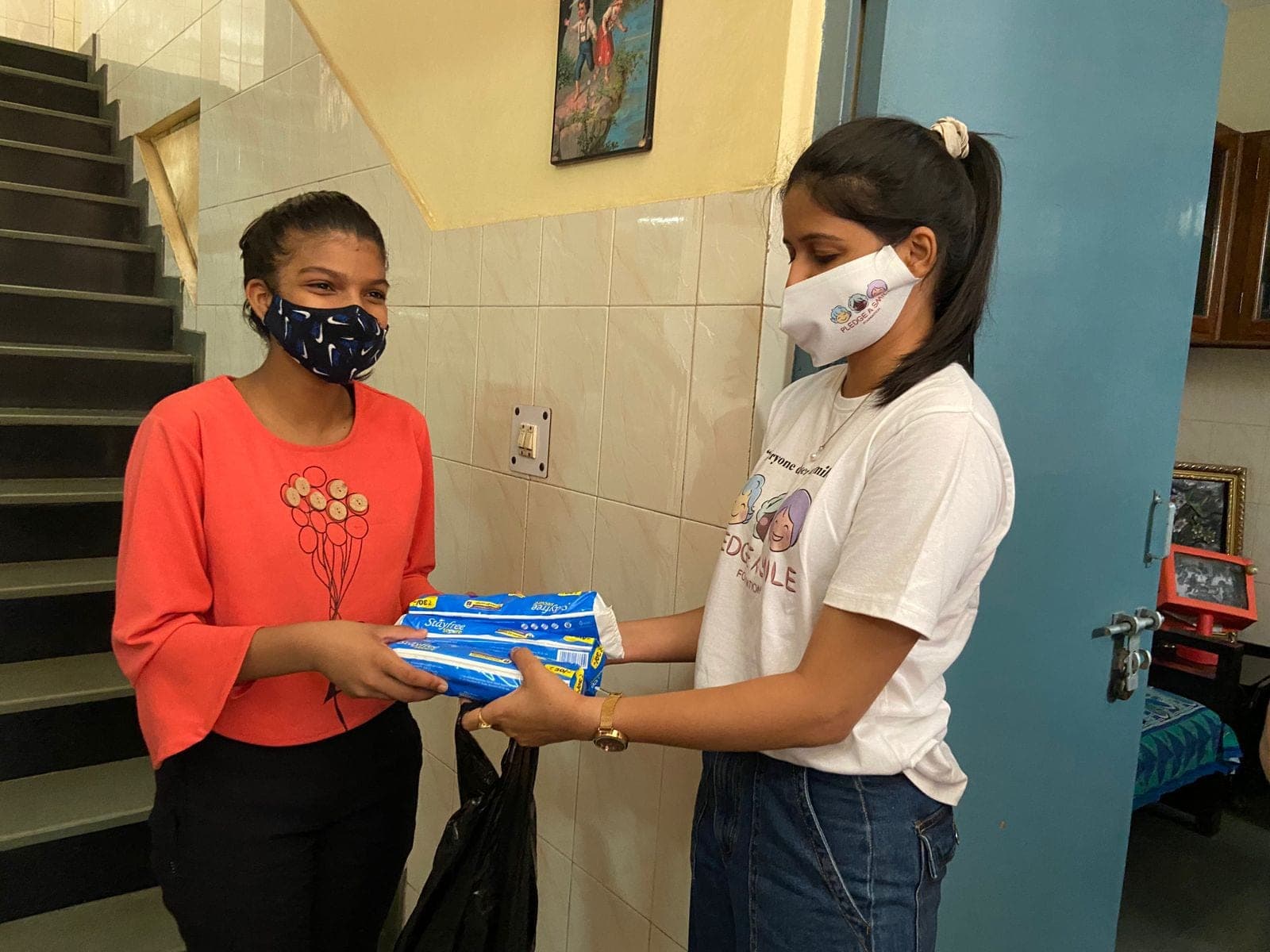 A girl in an orange top receives pads from a volunteer wearing a Pledge A Smile branded mask.