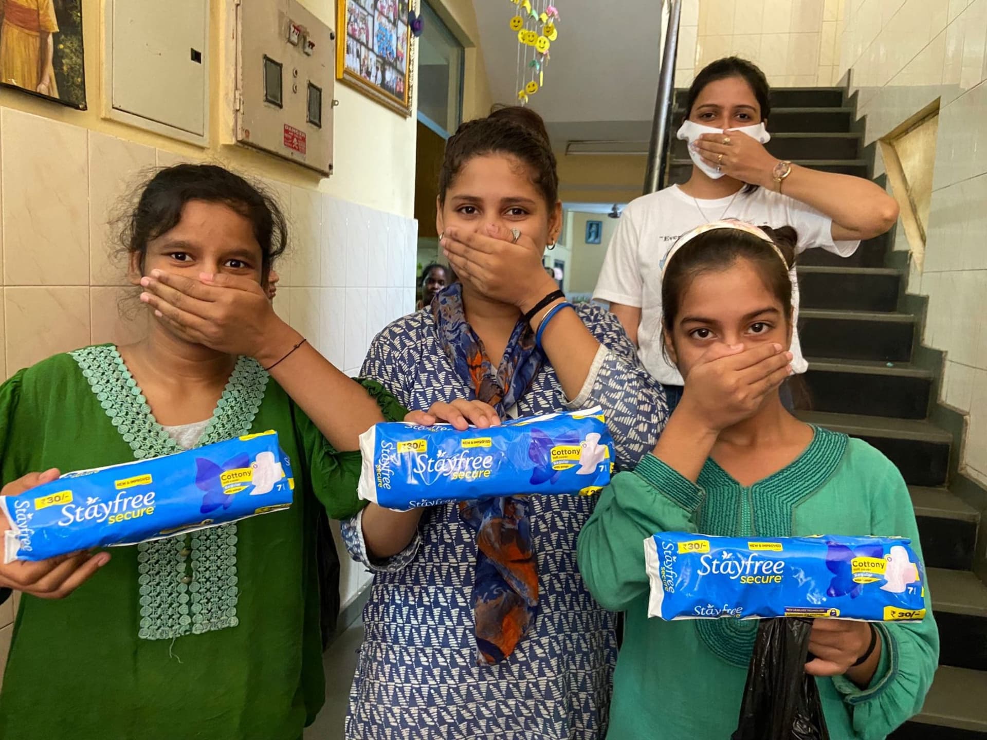 Three girls hold sanitary pads with their hands over their mouths