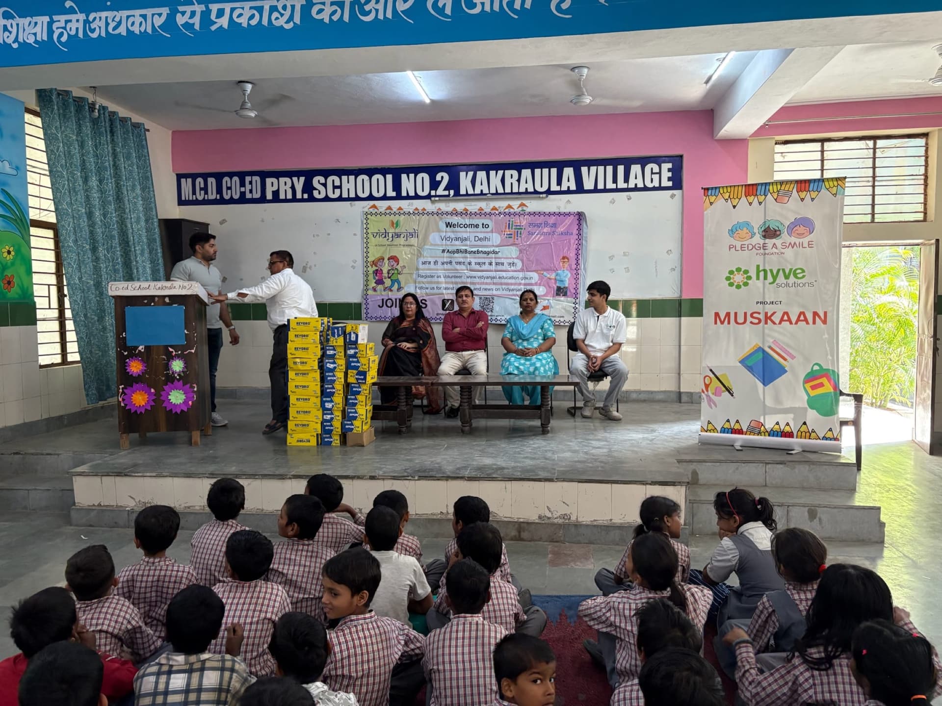 Children seated, watching a Muskaan distribution ceremony on a classroom stage