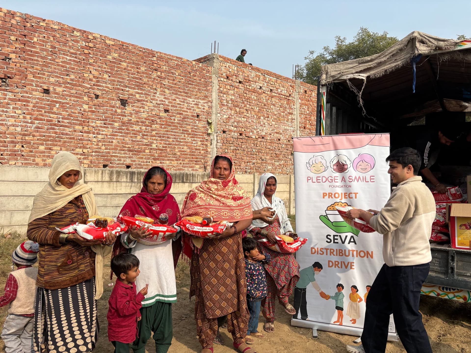 A crowd receives atta ration bags at a Project Seva drive by a brick wall